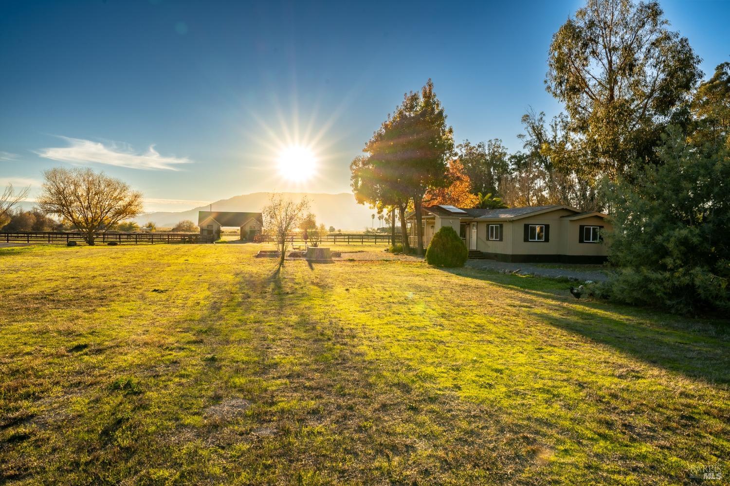 Bathed in the golden light of a setting sun, this modest ranch-style home sits peacefully in a sprawling field. The surrounding trees and distant mountains create a serene backdrop for this quiet country property.