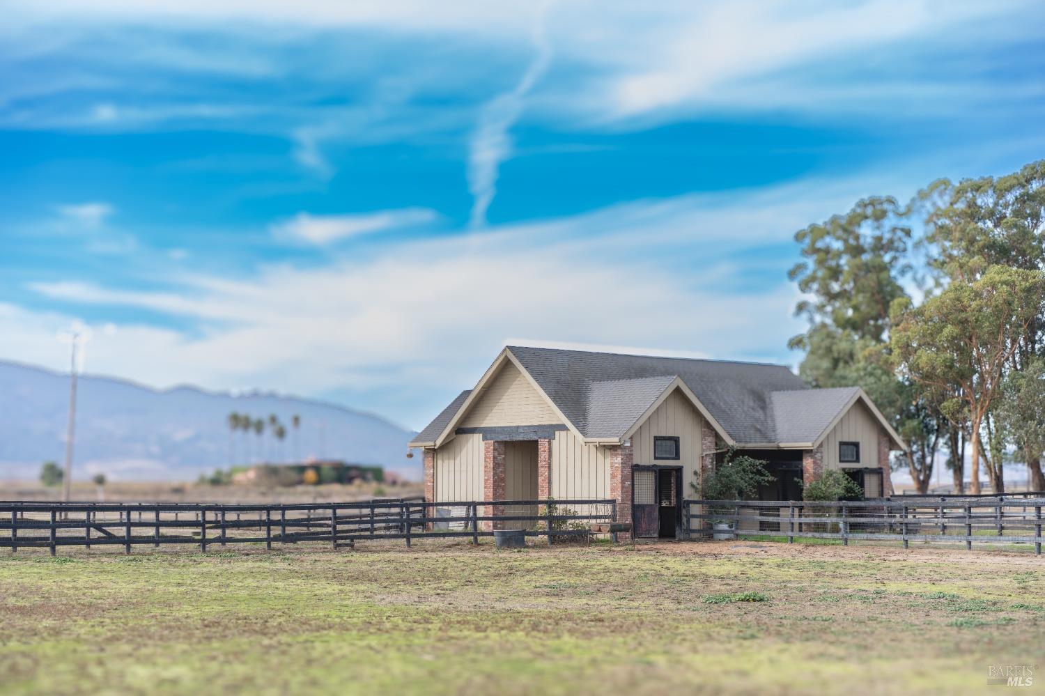 A rustic barn sits on a sprawling ranch, surrounded by lush green fields and a clear blue sky. The barn's weathered wood and brick construction add to its charm.