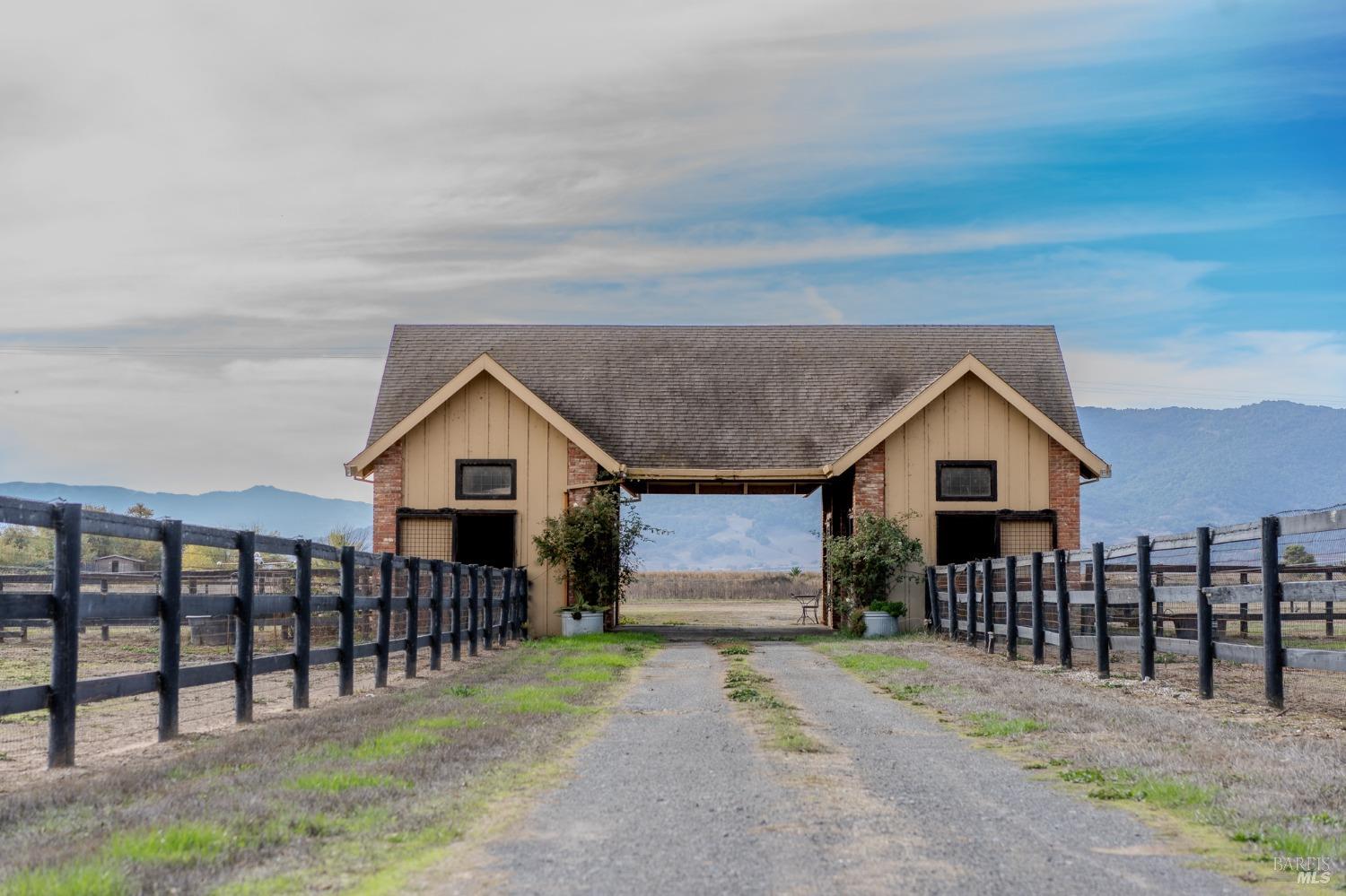 A classic stable, with its warm wood siding and sturdy brick foundation, stands proudly against a backdrop of rolling hills and a bright blue sky. The black fencing and expansive paddock complete the quintessential equestrian scene.