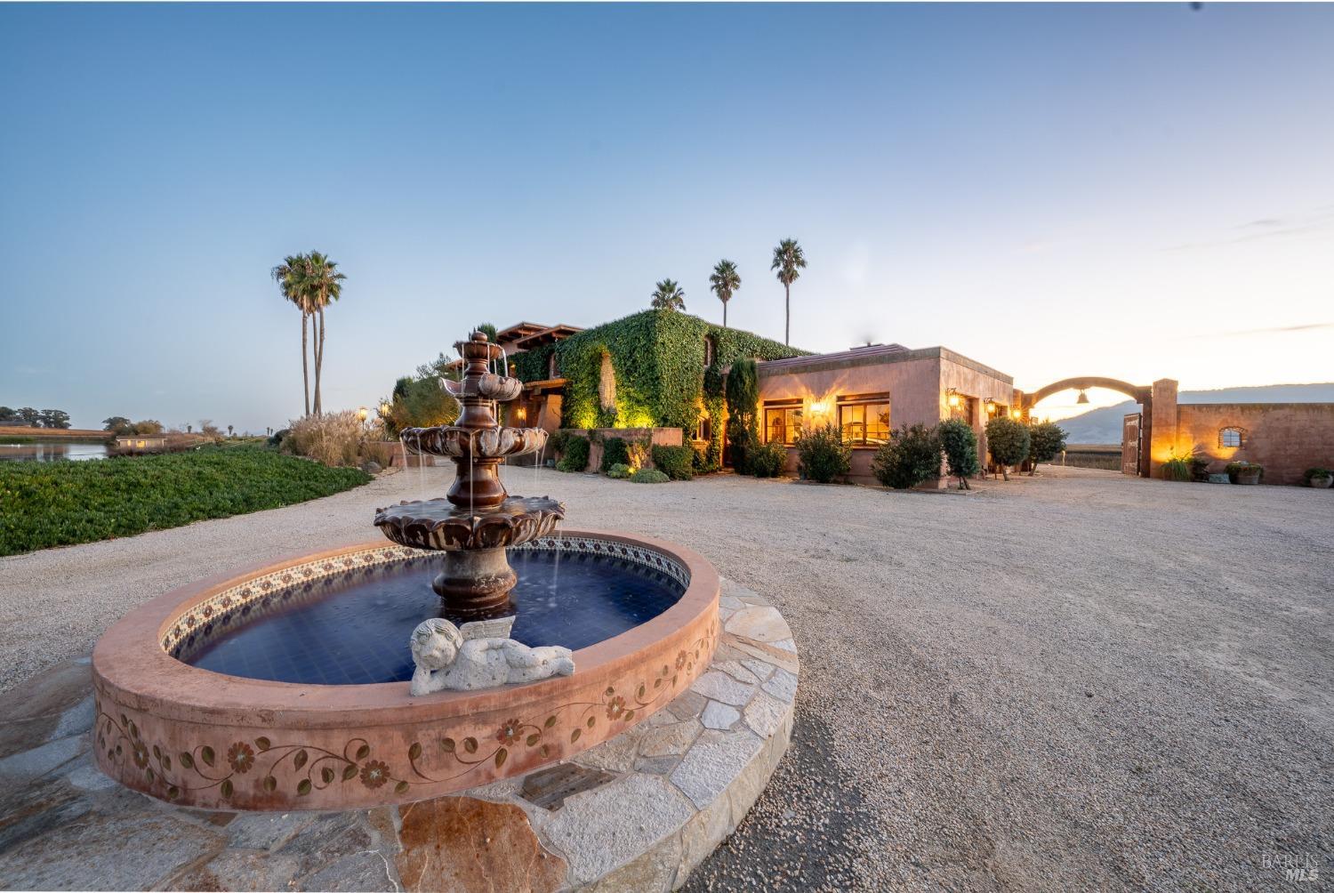 A beautiful fountain with a cherub statue in the center, surrounded by lush greenery and a grand Spanish-style mansion. The setting sun casts a warm glow on the scene, creating a romantic and idyllic atmosphere.