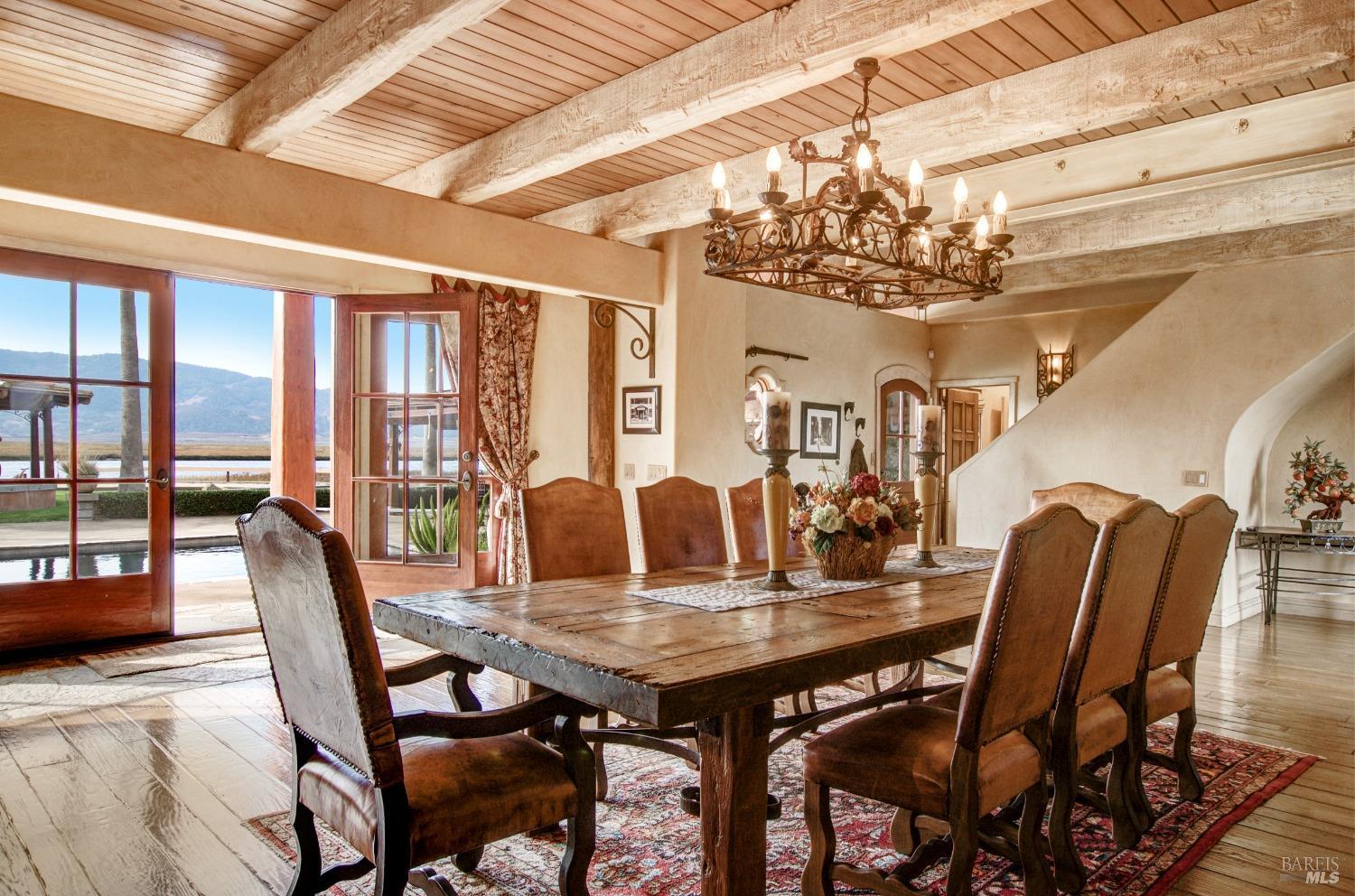 This elegant dining room boasts a rustic ambiance with its exposed wood beam ceiling and expansive French doors that open to a scenic waterfront view. The sturdy wooden dining table and leather chairs create a sense of warmth and invite comfortable gatherings.