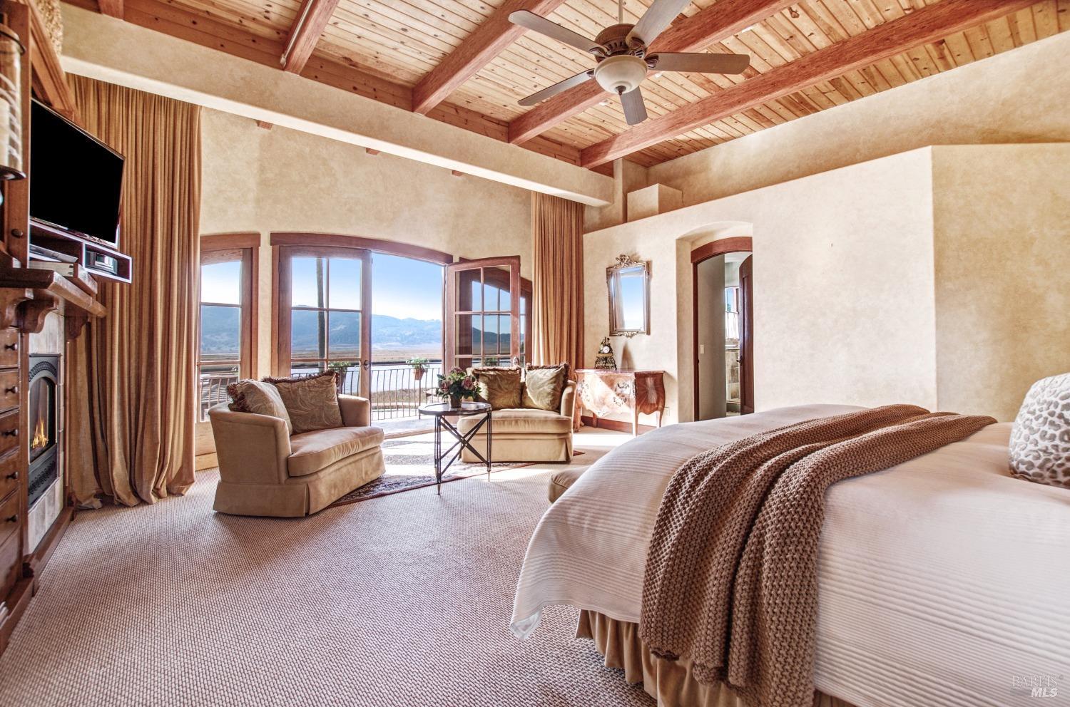 This luxurious bedroom features a cozy sitting area with a fireplace and breathtaking views, creating a serene and inviting retreat. The high ceilings with exposed beams, plush carpeting, and elegant furnishings add to the room's overall sense of grandeur and comfort.