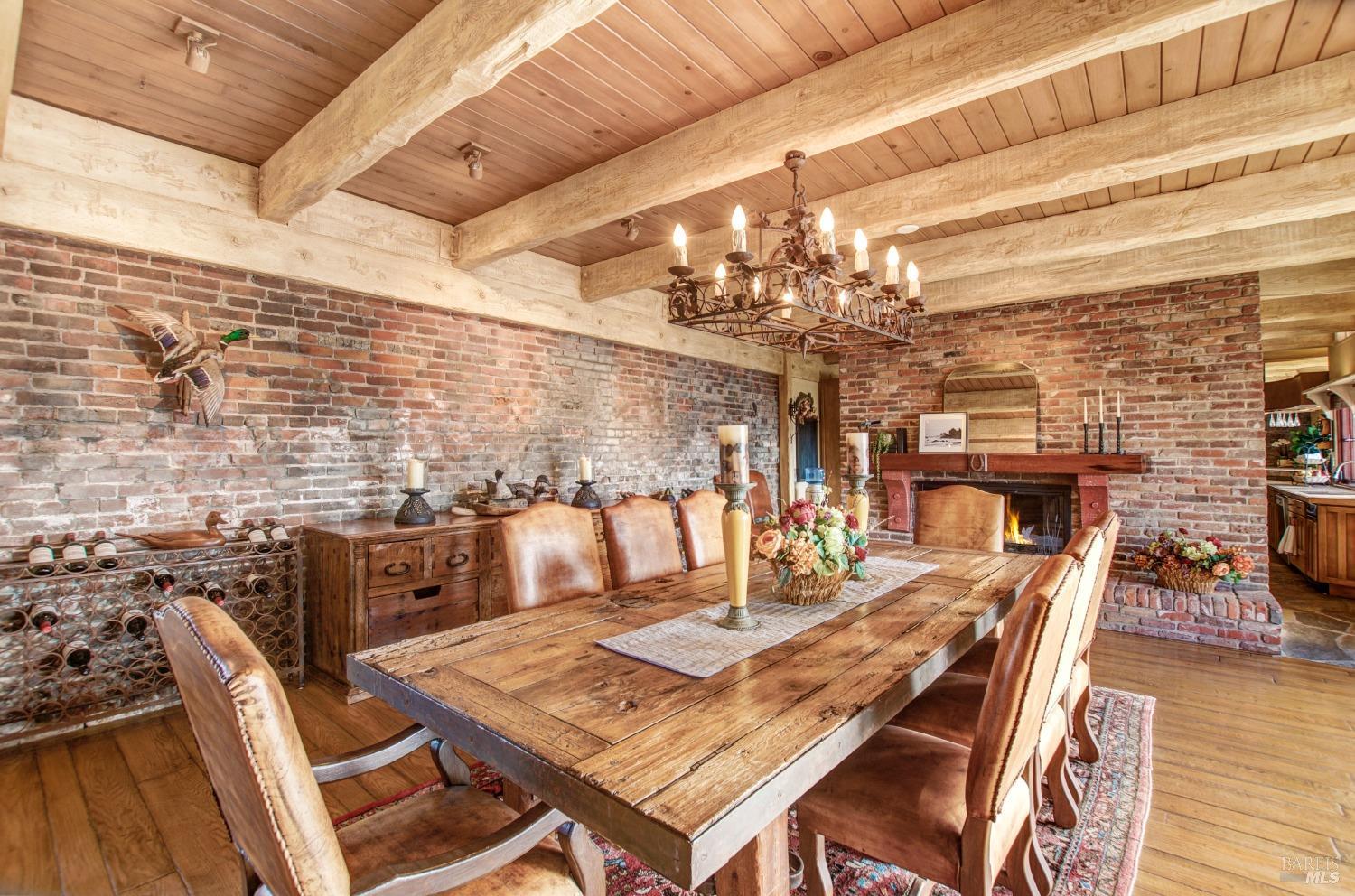 This grand dining room features rustic charm with exposed brick walls, wood beamed ceilings, and a large, rugged dining table. The chandelier and fireplace add a touch of elegance to the space, creating a warm and inviting atmosphere for gatherings.