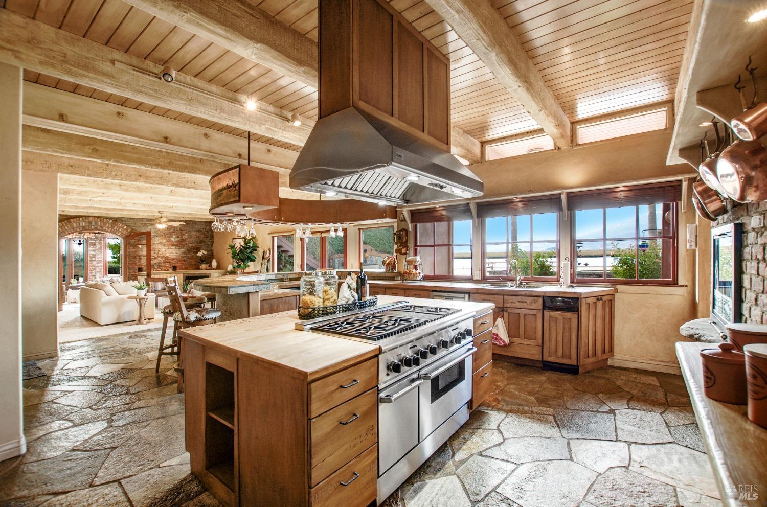 A rustic and spacious kitchen features a large island with a professional-grade range and ample counter space, perfect for culinary endeavors. The exposed beams, stone flooring, and large windows with scenic views create a warm and inviting atmosphere for cooking and entertaining.