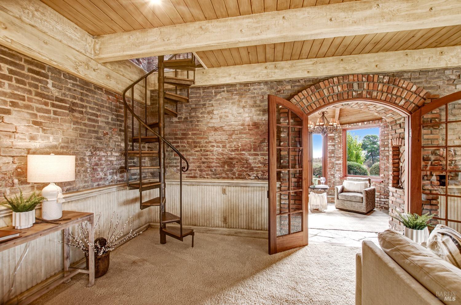 This room seamlessly blends rustic and industrial elements with its exposed brick walls, wood-beamed ceiling, and a striking spiral staircase. French doors open to a charming brick patio, further enhancing the appeal of this unique space.