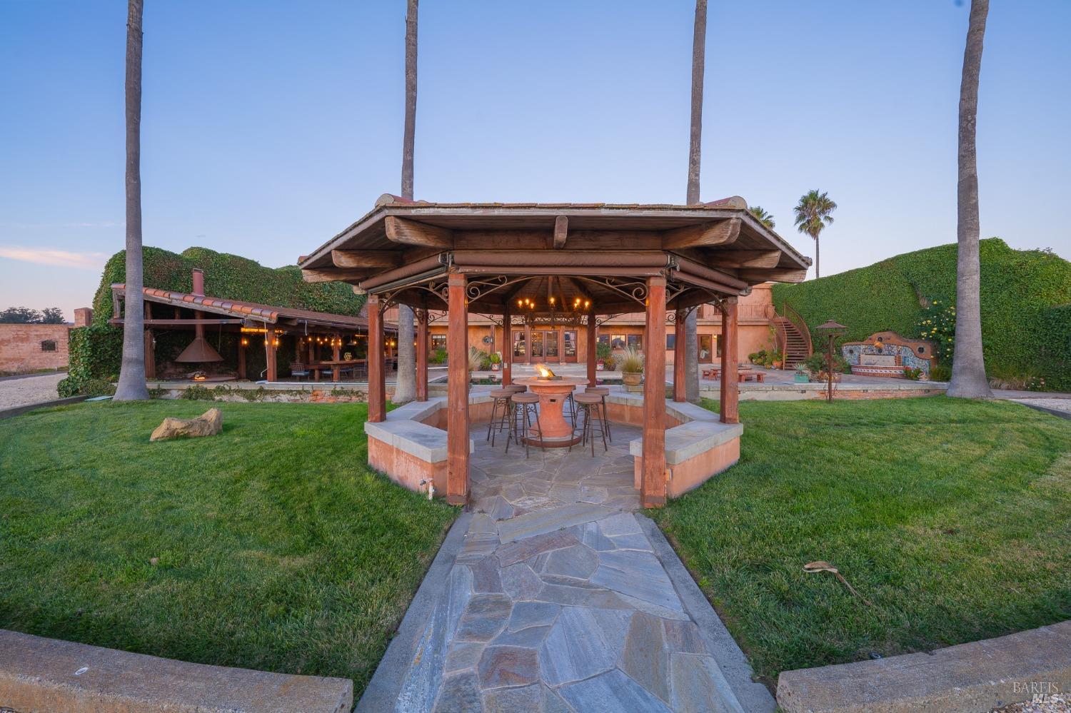 A welcoming gazebo with a cozy fire pit forms the centerpiece of this outdoor living space, inviting relaxation and socializing. The surrounding lush lawn, palm trees, and ivy-covered buildings create a tranquil and inviting atmosphere.