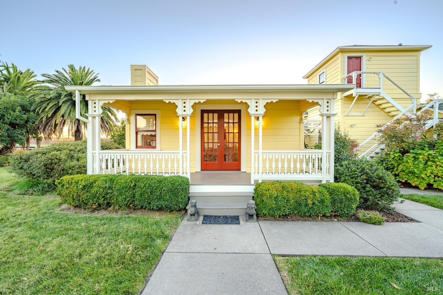 This charming yellow cottage features a welcoming front porch with classic white railings and a set of inviting red double doors. The well-maintained landscaping and quaint architecture give the home a cozy and inviting curb appeal.