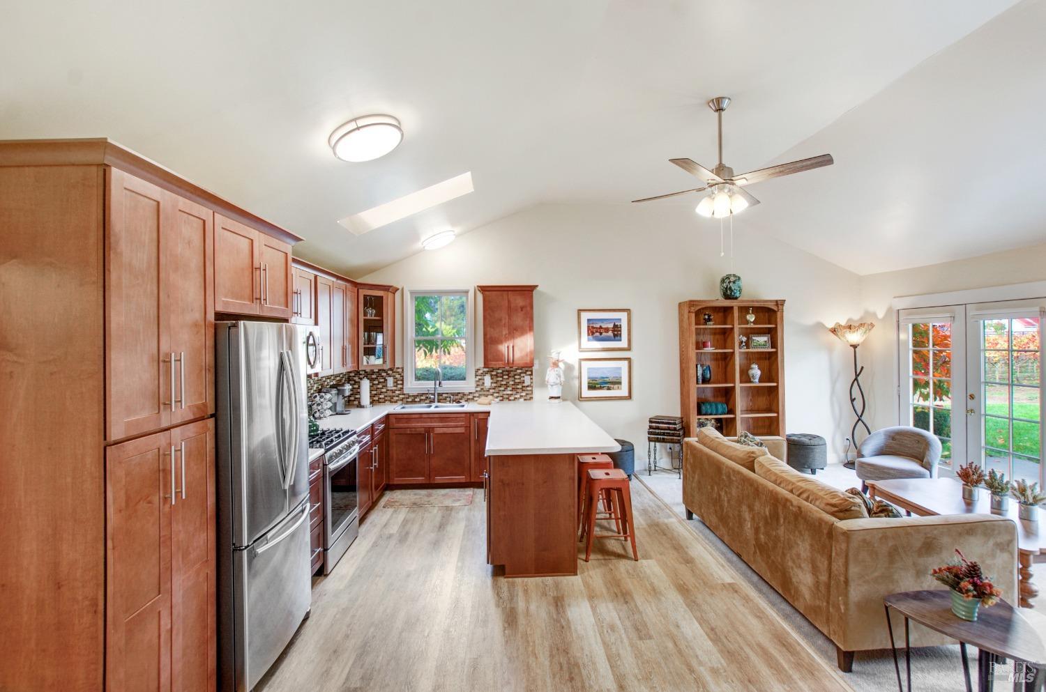 This is a modern open-plan living space with a combined kitchen, dining, and living area. The kitchen features warm wood cabinets, stainless steel appliances, and a breakfast bar, while the living area includes a comfortable sofa and access to the backyard.