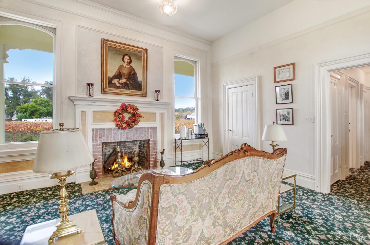 A cozy sitting room exudes classic charm with its ornate fireplace, antique furnishings, and decorative rug. Large windows offer a picturesque view, adding to the room's inviting ambiance.