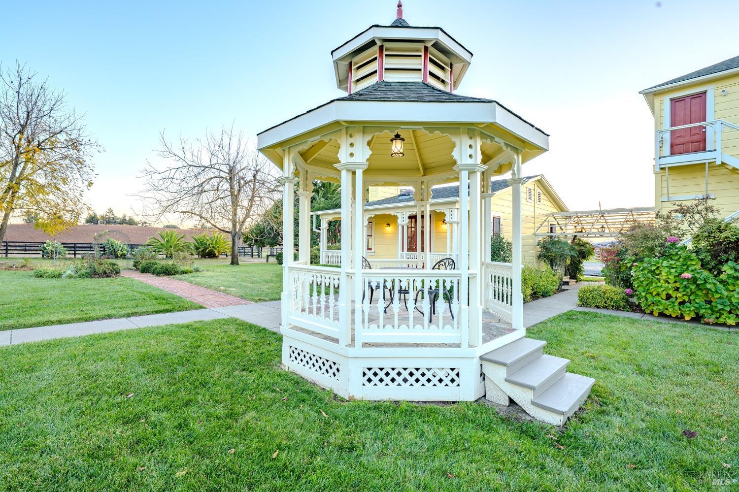 A classic white gazebo with a charming red roof provides a picturesque focal point in a serene garden setting, offering a peaceful spot for relaxation or contemplation. The surrounding lush lawn, vibrant flowers, and pathways invite leisurely strolls and enjoyment of the outdoor space.