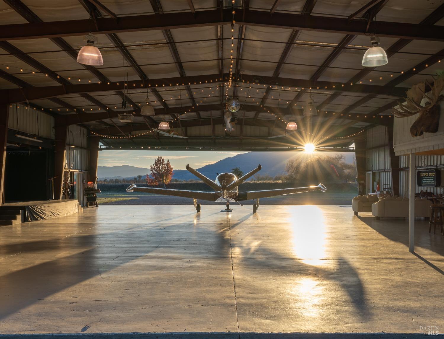 This multi-purpose hangar seamlessly blends functionality with a touch of magic. With a private plane at its center, bathed in the golden light of the rising sun, it also boasts a stage, suggesting it's used for events and gatherings, making it a truly unique and versatile space.