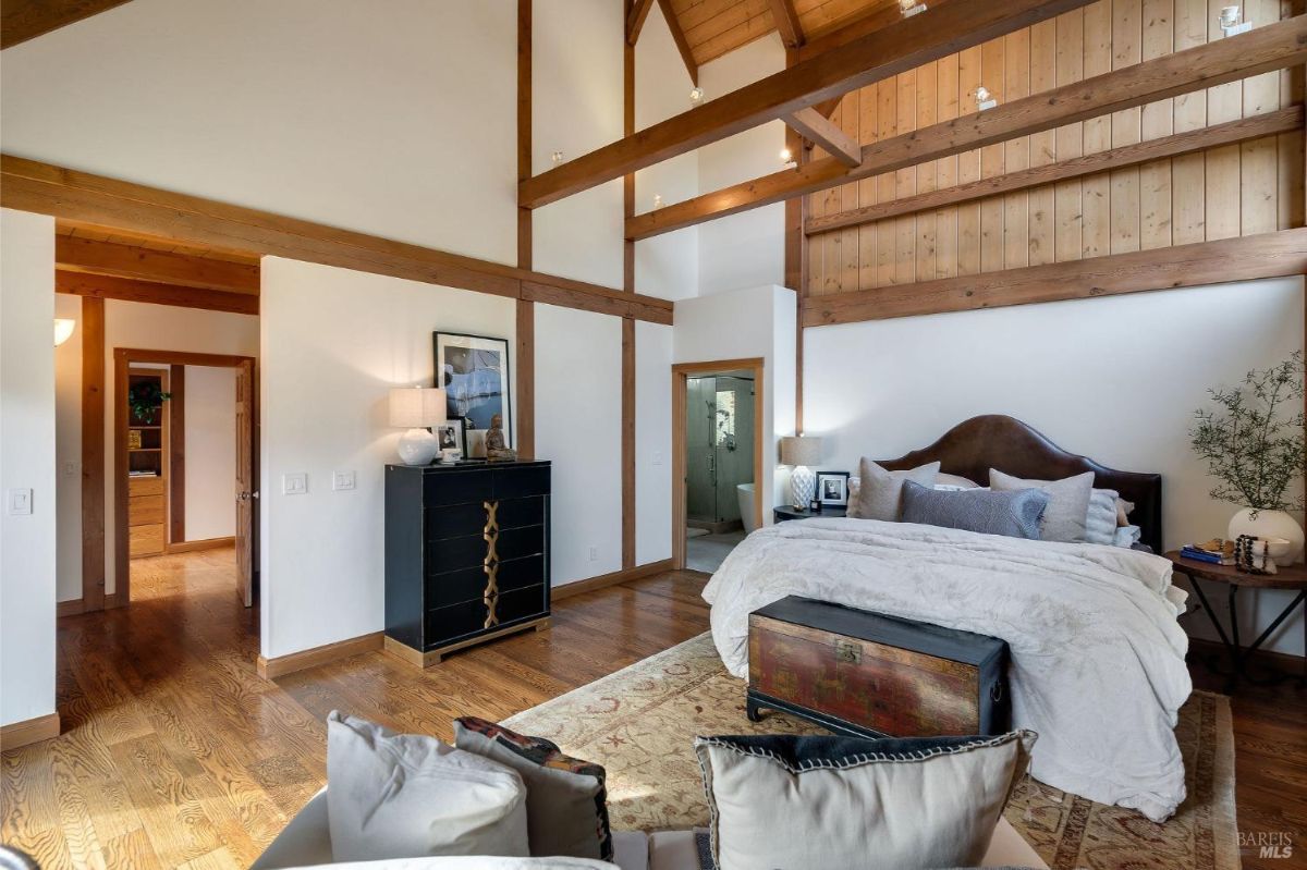 Spacious bedroom with high wood ceilings, decorative beams, and a connected bathroom visible through an open door.