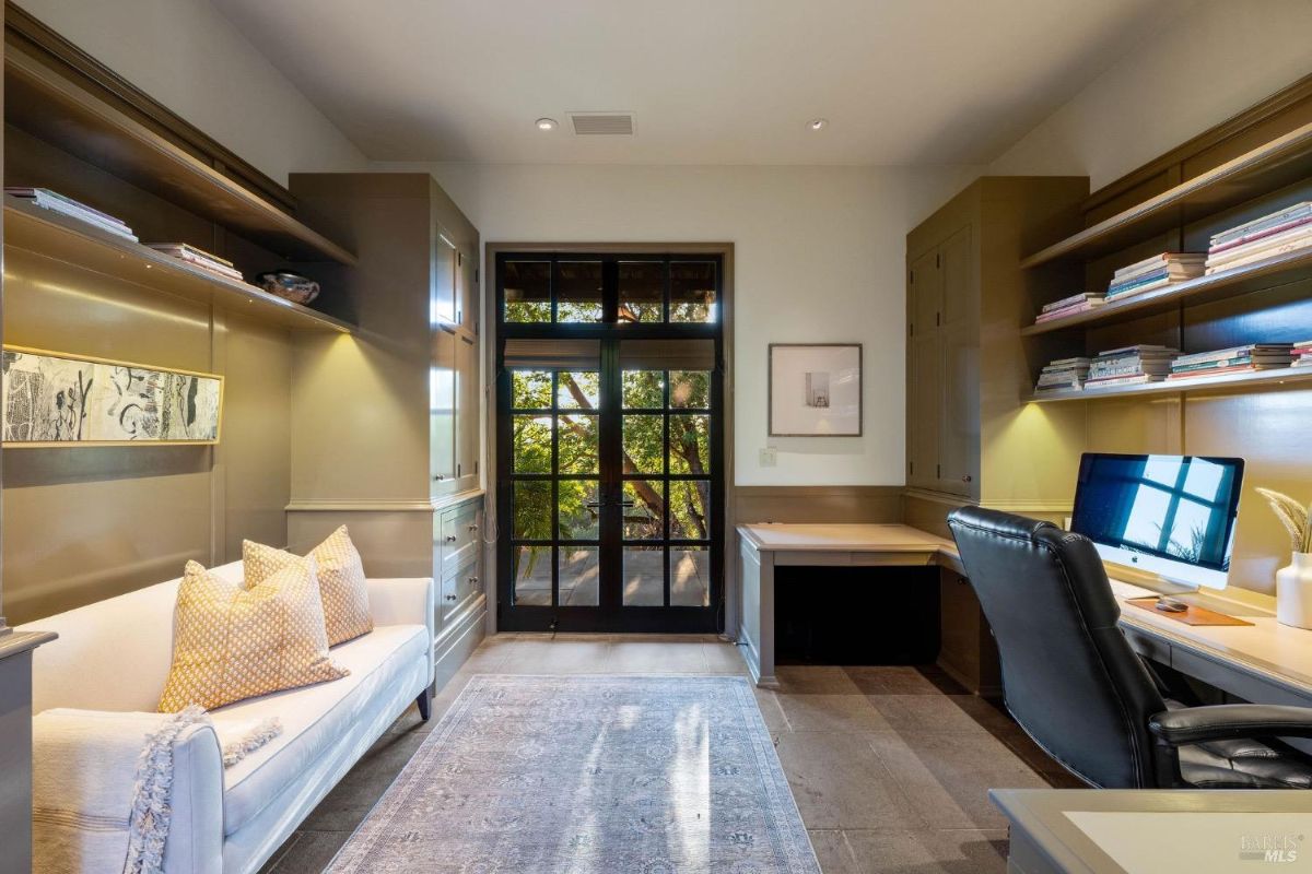 Home office with built-in shelves, a desk, and a sofa, with a view of the outdoors through double glass doors.