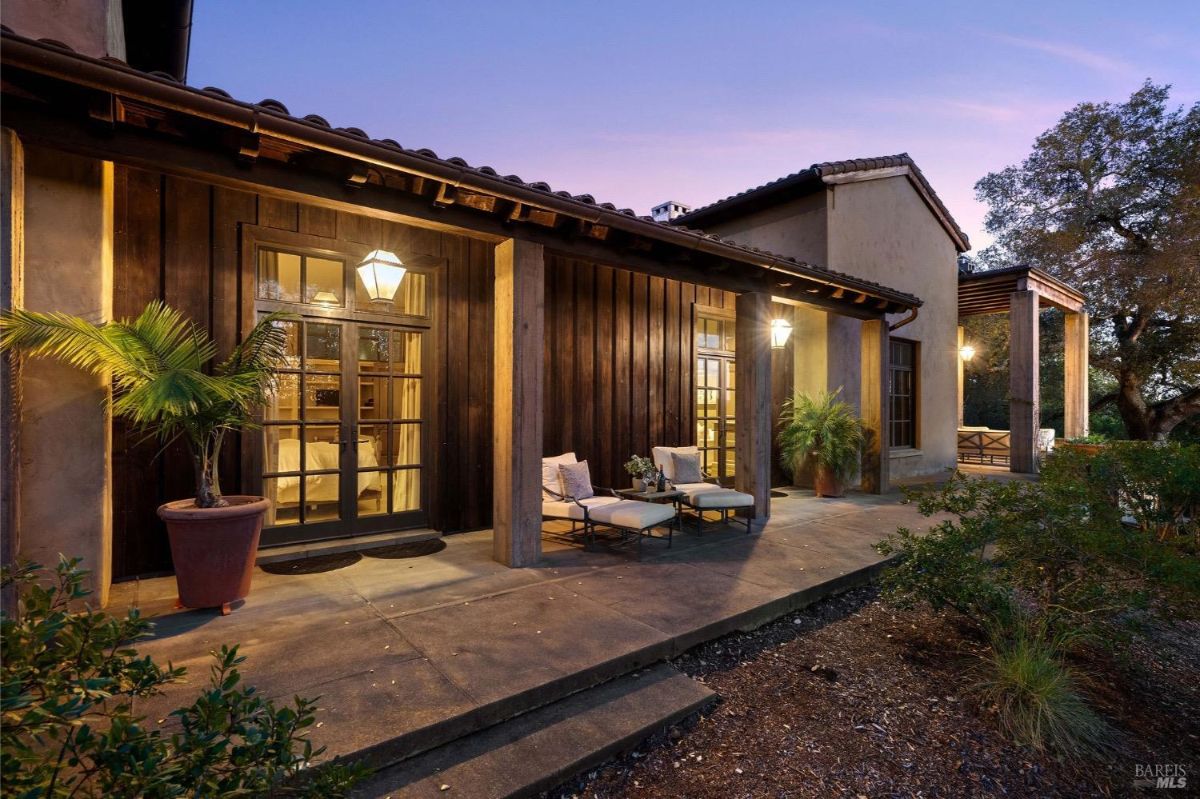 Exterior of a house at dusk, featuring a patio with seating and illuminated lanterns near wooden and stucco walls.