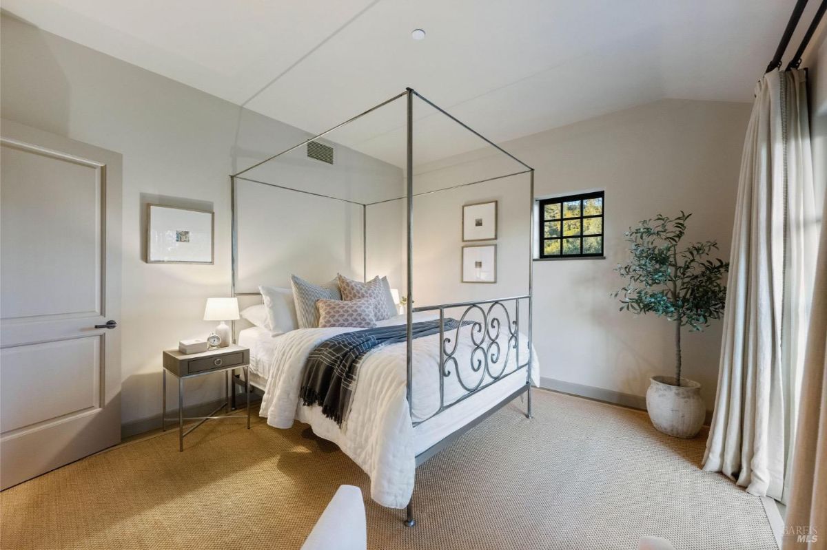 Bedroom with a decorative iron canopy bed, beige carpet, and a large potted plant near the window.