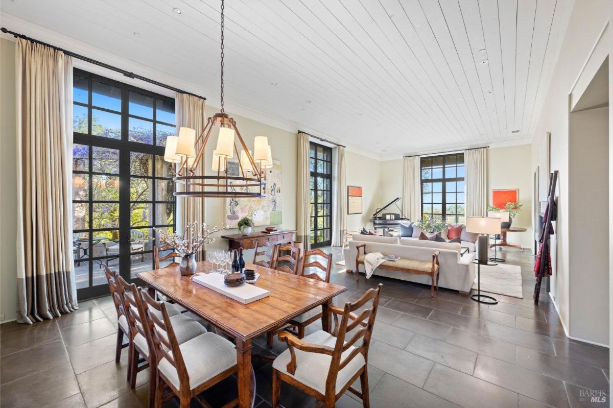 Dining area with a wooden table and chairs beneath a chandelier, adjacent to a living space with large windows.