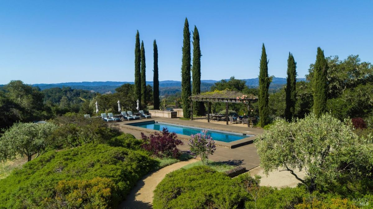 Landscape view of the pool area with a pathway through lush vegetation and cypress trees in the background.