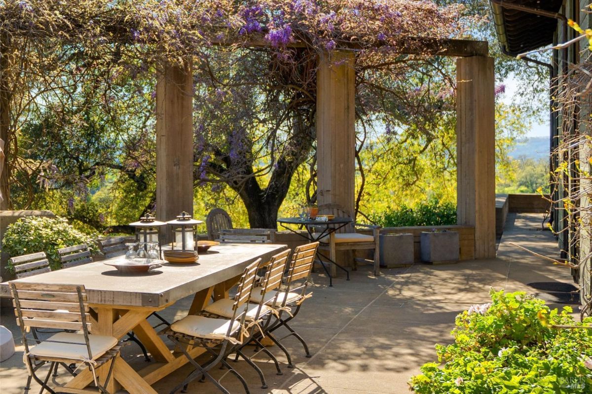 Outdoor dining area under a pergola with vines and trees providing shade.