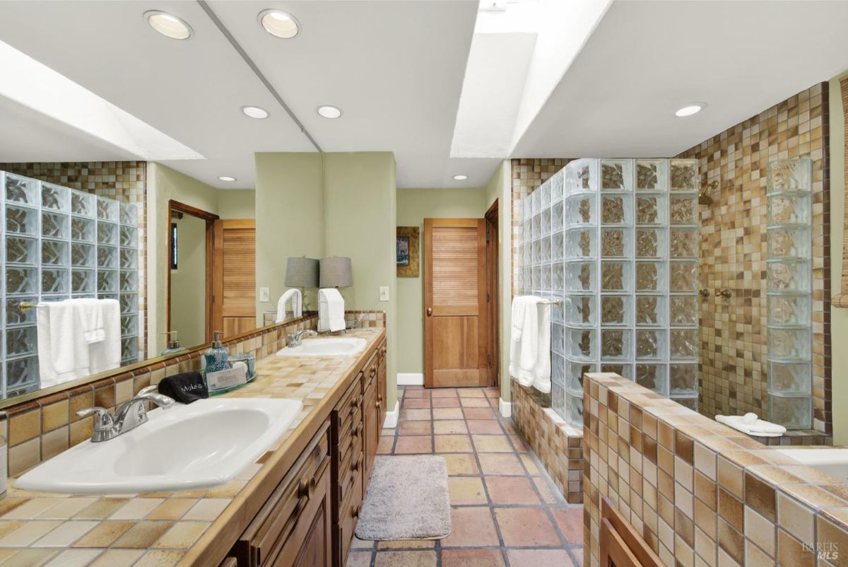 The bathroom has two sinks, a large mirror, a glass block shower enclosure, and tiled floors.