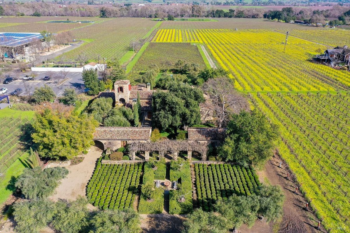 Aerial view of a property surrounded by vineyards. The property includes a building with a tile roof, a parking area, and adjacent farmland with rows of vines extending into the distance.