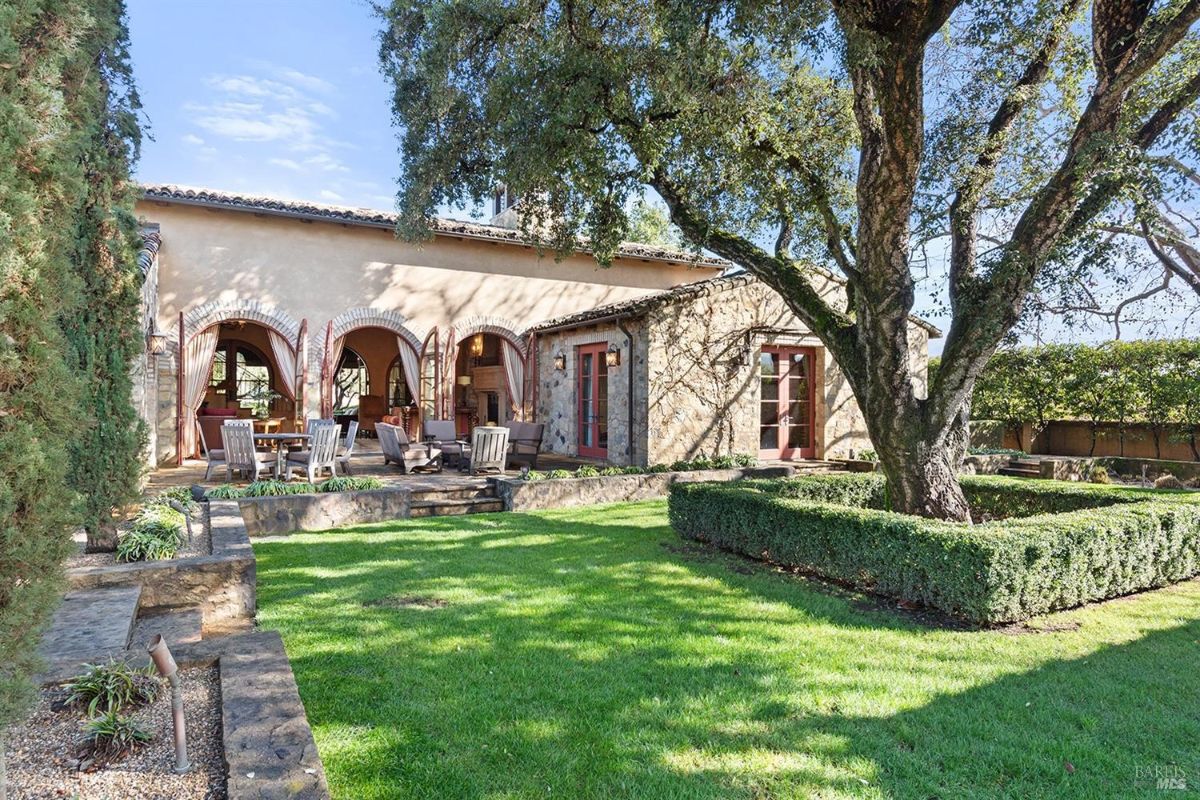 This picturesque backyard oasis features a charming stone house with arched doorways and a welcoming patio area. A majestic tree provides shade and a sense of tranquility, while the manicured lawn and surrounding greenery create a peaceful and inviting atmosphere for relaxation and outdoor enjoyment.