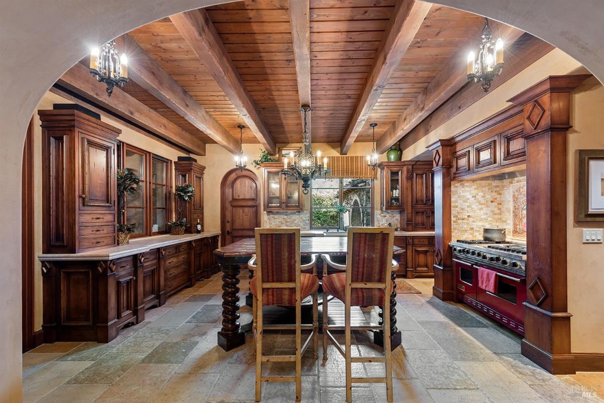 A kitchen with exposed wooden beams, wood cabinetry, and a central island with seating. Includes a large stove with a decorative tiled backsplash. 