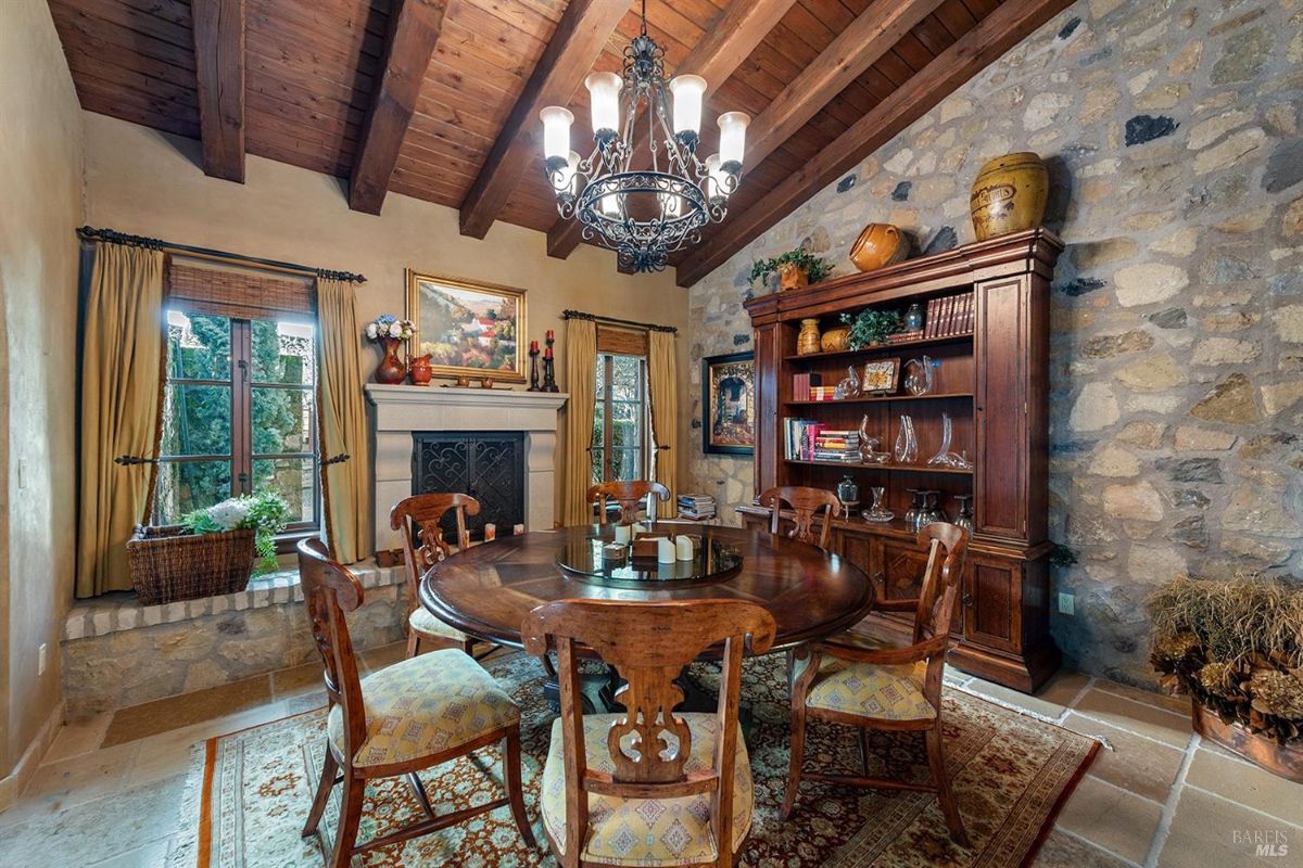 Another view of the dining room showing the same circular wooden table and chairs, fireplace, and wooden cabinet with books and decor. 