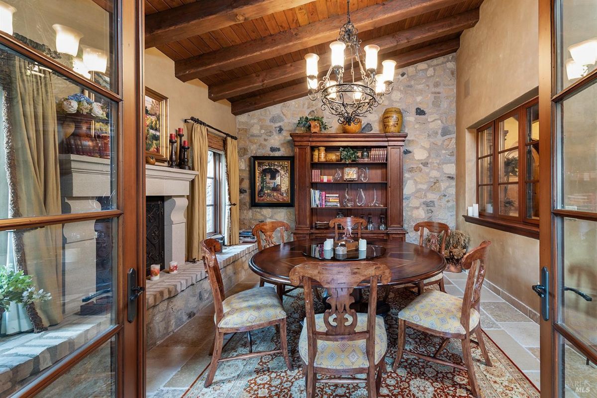  A dining room with a circular wooden table, six chairs, a fireplace with a mantel, and a wooden cabinet against a stone wall.