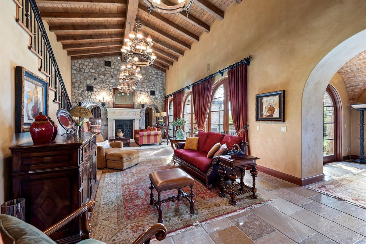 Interior view of a large living room with exposed wooden beams, stone walls, and chandeliers. The room includes a fireplace, furniture such as sofas and chairs, and large arched windows with curtains.