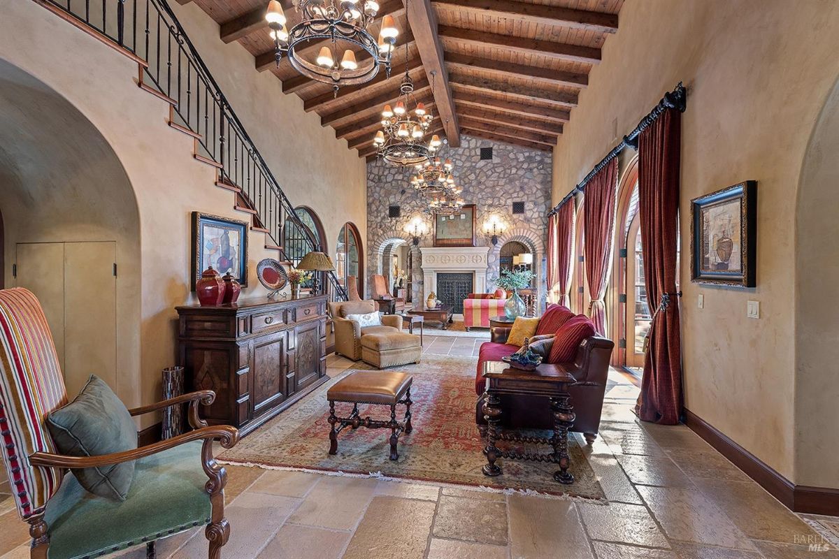 A full view of the large room showcasing the fireplace, seating arrangements, stone walls, and high ceiling with exposed beams and chandeliers. The room has a central rug and decorative elements like paintings and vases.