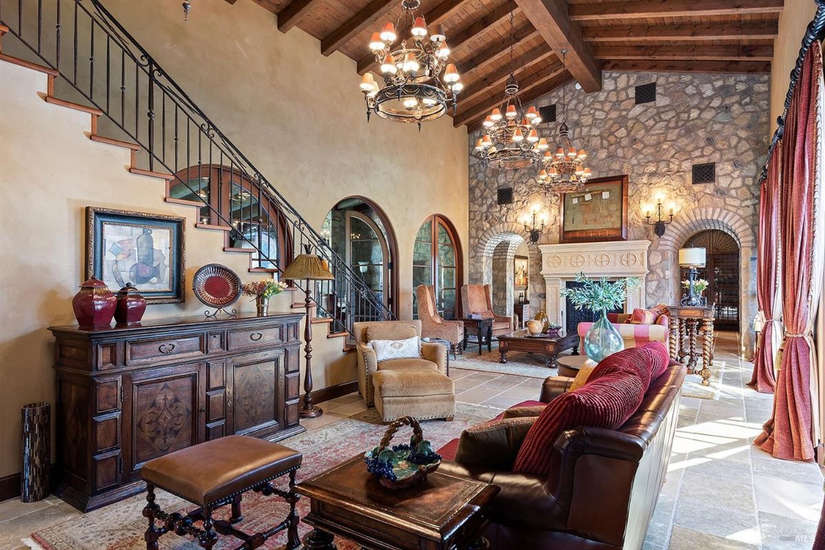 A view from the end of the room, emphasizing the fireplace, arched doorways, and a seating area with a sofa and armchairs. The room has symmetrical features, including chandeliers and window placement.