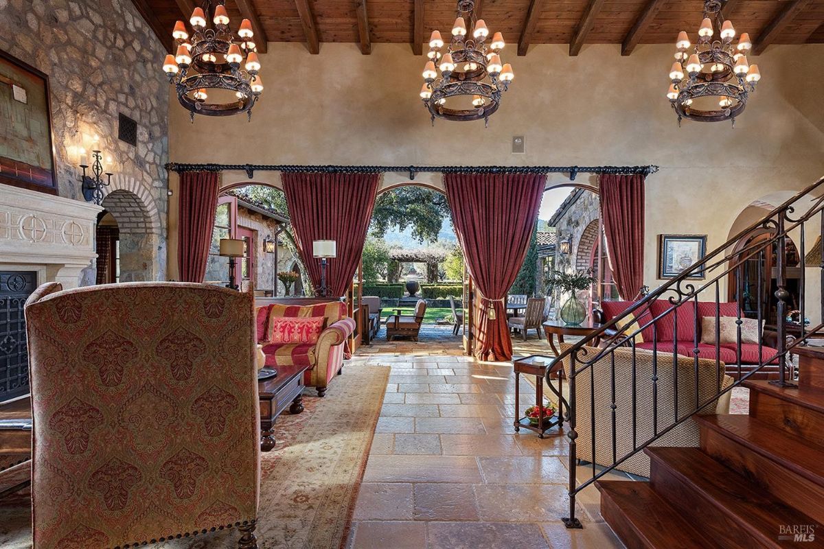 Interior view of a large room with exposed wooden beams, chandeliers, stone walls, and large arched windows. The room features a fireplace, chairs, a sofa, and an open view of an outdoor courtyard through arched doorways.