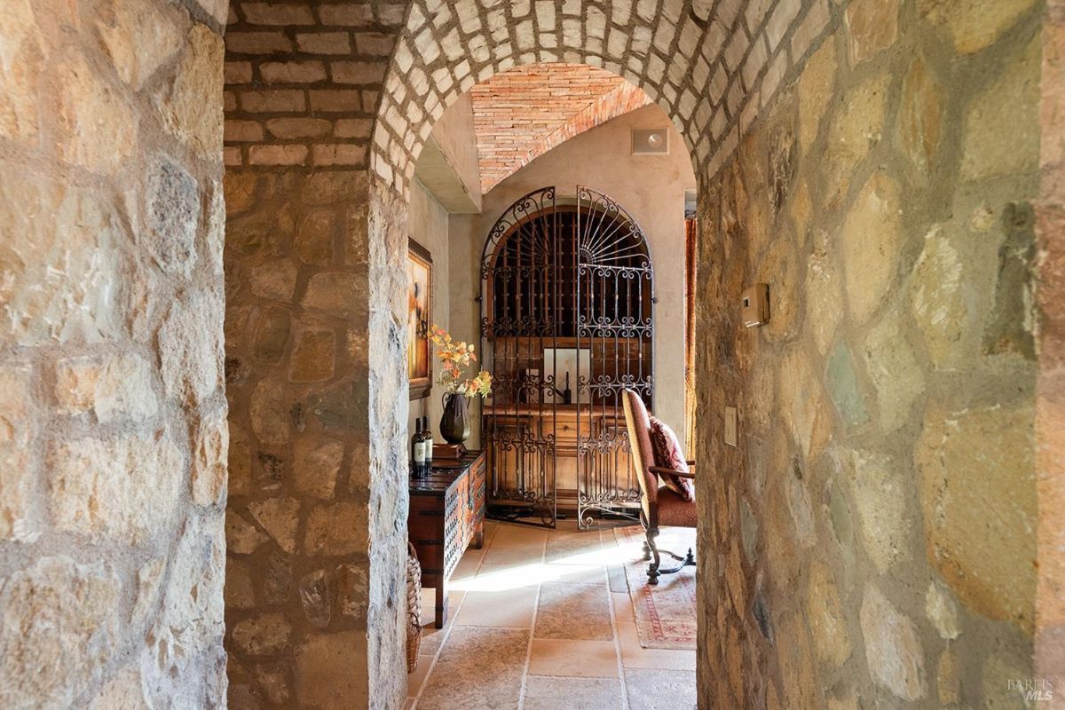 A hallway with stone walls and an arched ceiling. A decorative wrought iron gate is visible, along with a wooden sideboard and a chair.