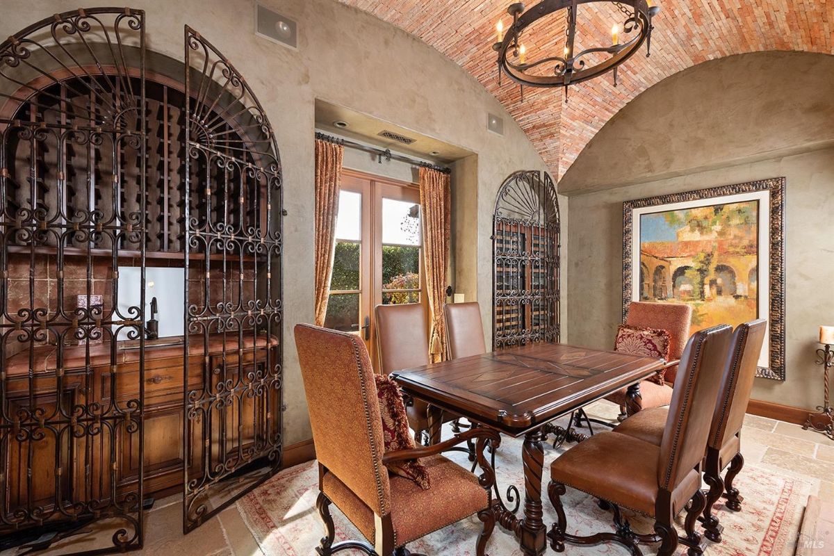 A dining room with a brick vaulted ceiling, wrought iron wine racks, and a wooden table surrounded by chairs. A painting and arched windows are visible.