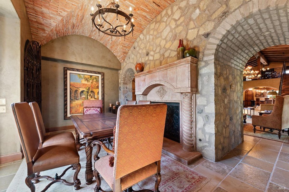  A dining area with a stone fireplace, a wooden table, and chairs. The ceiling features a brick design with a chandelier. An arched doorway connects to another section of the property.