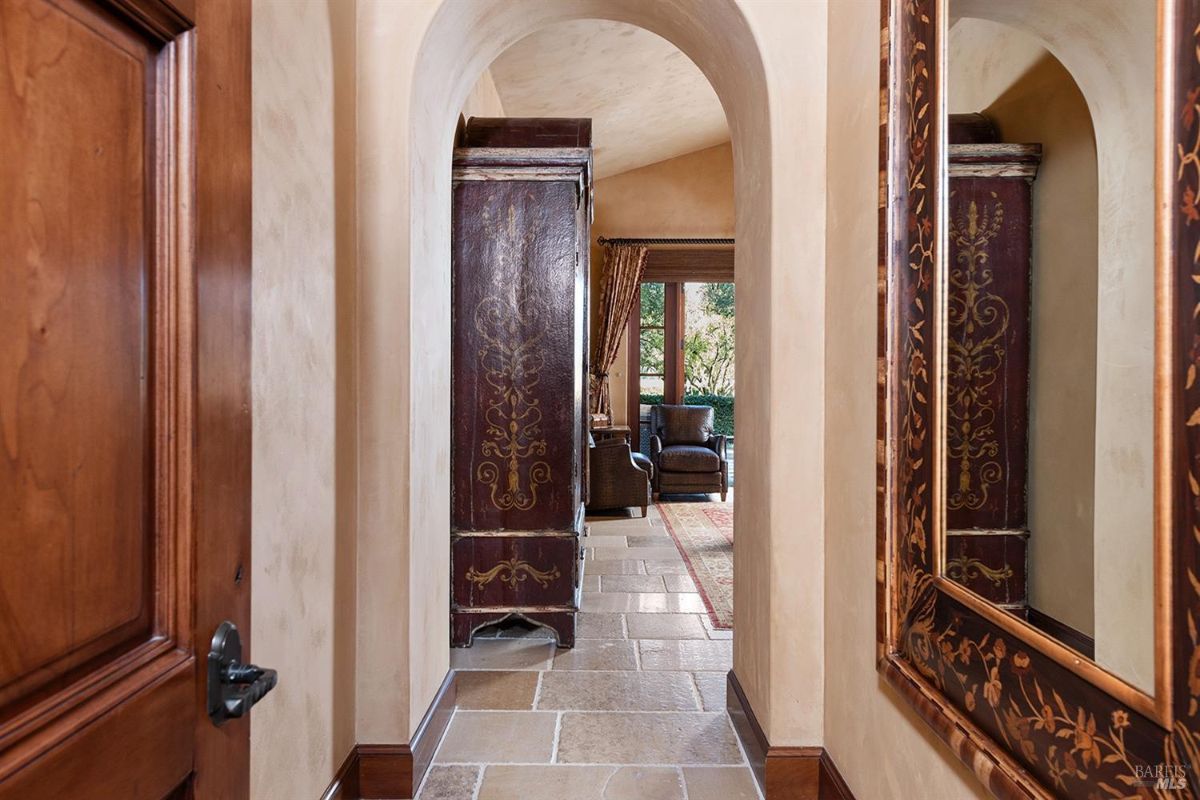 A hallway with arched doorways leading to a room. A mirror is mounted on the wall, and a decorative wardrobe is in view.