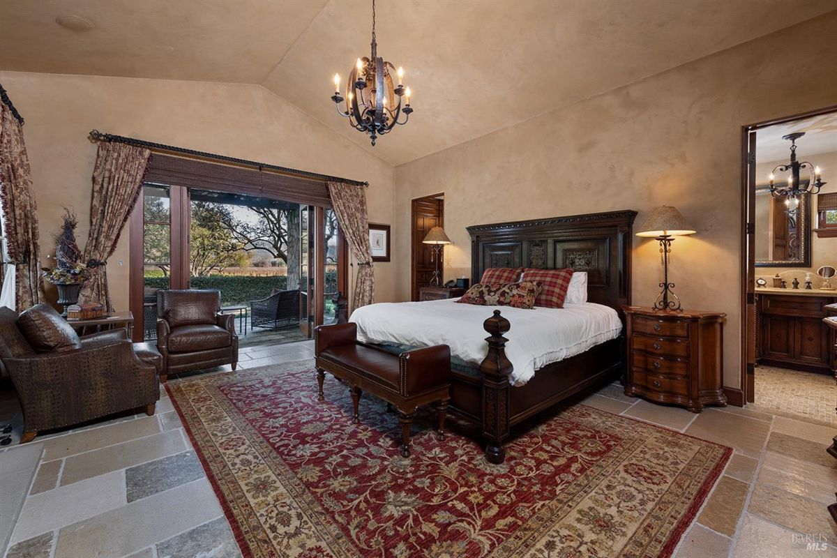 A bedroom with a bed, chairs, a rug, and large glass doors opening to a patio with outdoor furniture.