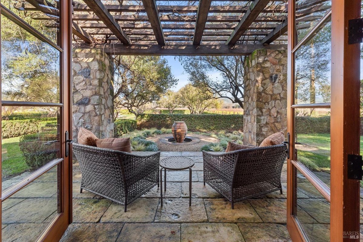 This inviting patio, sheltered by a rustic pergola, offers a tranquil space for relaxation and contemplation. Comfortable wicker chairs are perfectly positioned to enjoy the view of the garden, complete with a charming water feature and lush greenery. The open French doors suggest a seamless transition between indoor and outdoor living.