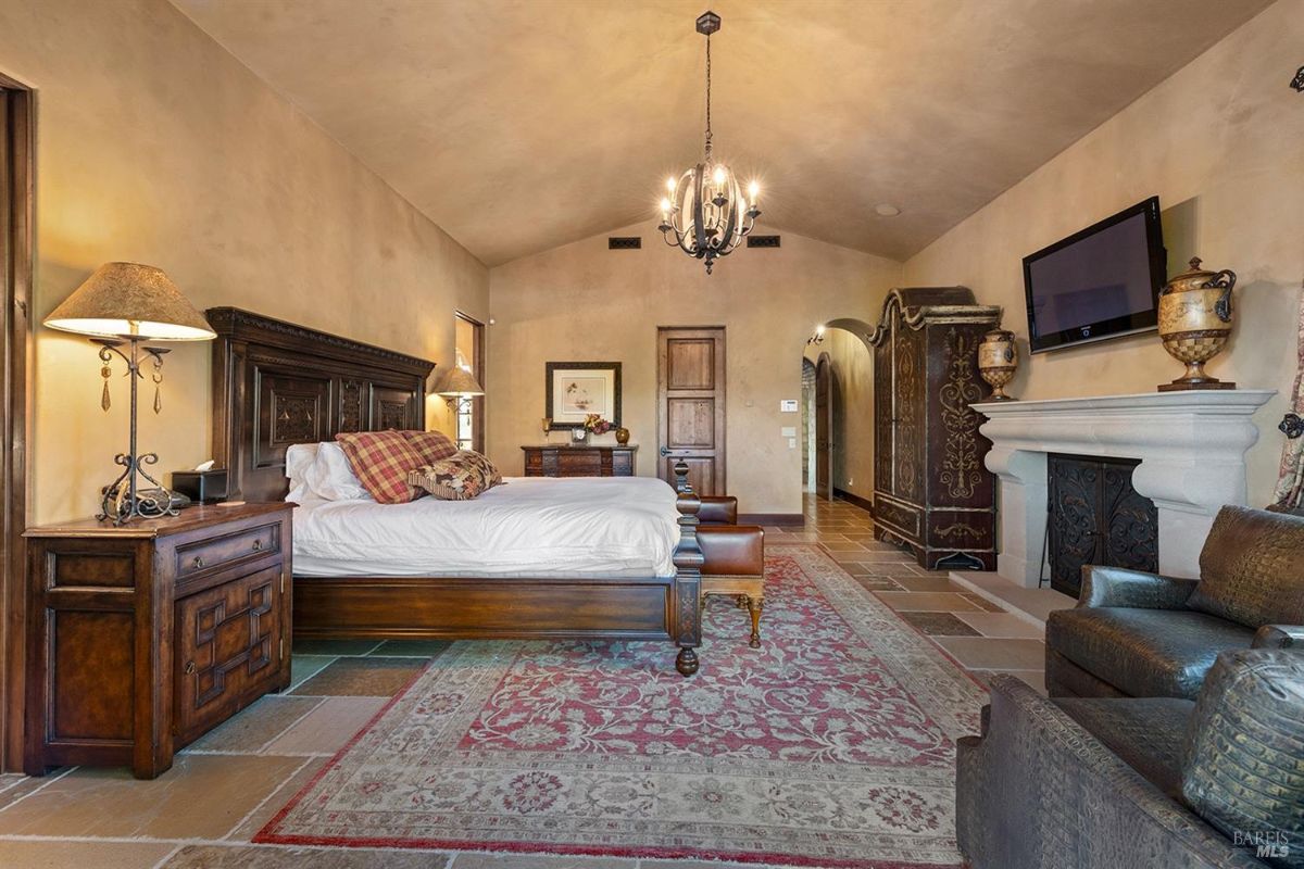 A bedroom with a bed in the center, nightstands on each side, a dresser, and a fireplace. A chandelier is hanging from the ceiling.