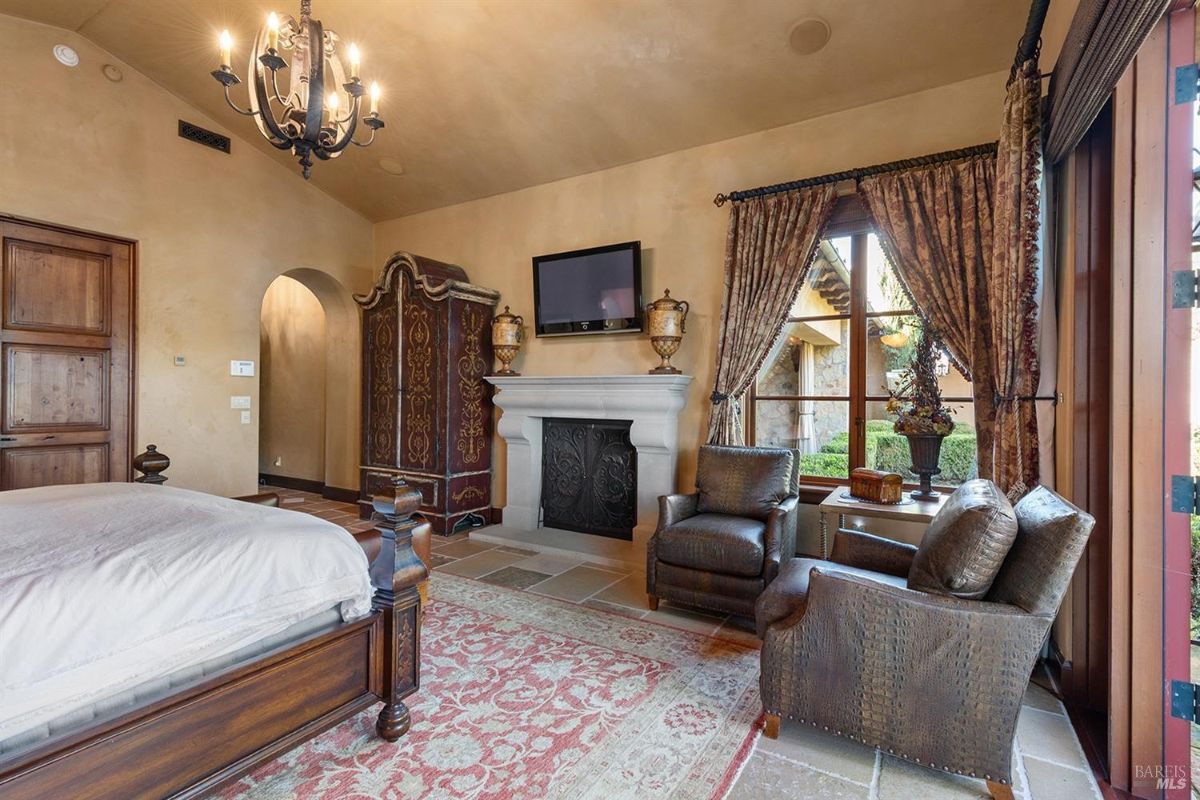 A bedroom with a bed, fireplace, and two armchairs. Curtains frame large windows leading to an outdoor view.