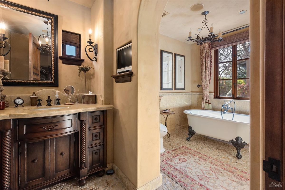 A bathroom with a freestanding bathtub, a sink with a vanity, and a mirror. There are framed windows with curtains, and the floor has small mosaic tiles.