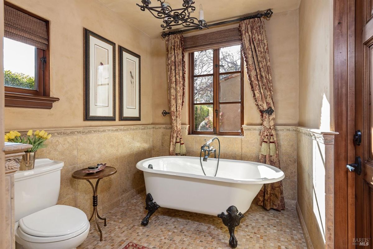 A bathroom with a freestanding clawfoot bathtub, tiled walls, a window with curtains, and a toilet on the left side. The floor is covered with mosaic tiles.