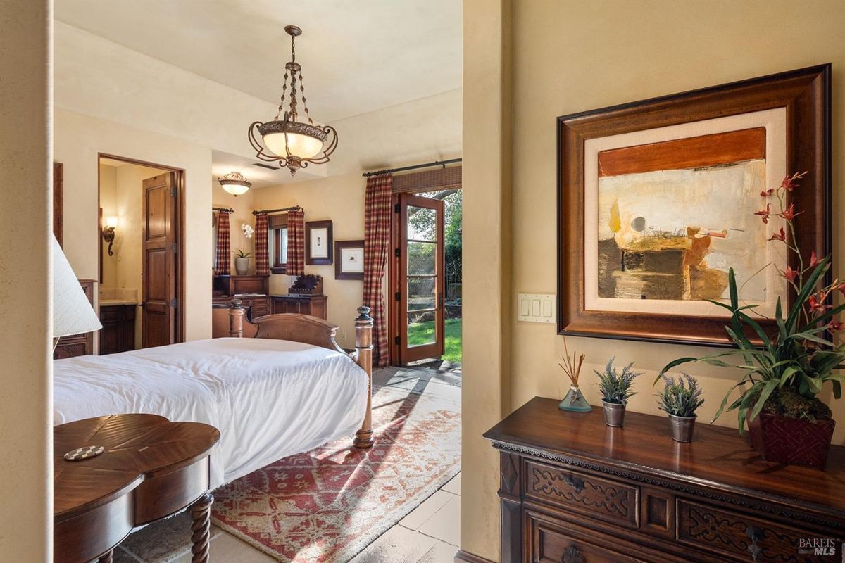  A bedroom with a wooden bed frame, a chandelier, and a side table. There is a painting and a plant on a dresser near the wall. A door leads to the outside with a view of greenery.