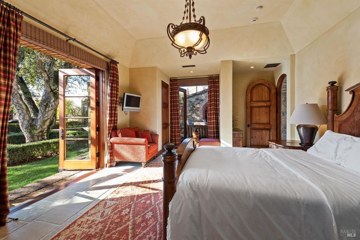 A bedroom with a wooden bed, an open door leading to a garden, a small red sofa, and a television mounted on the wall. There are plaid curtains on the windows and doors.