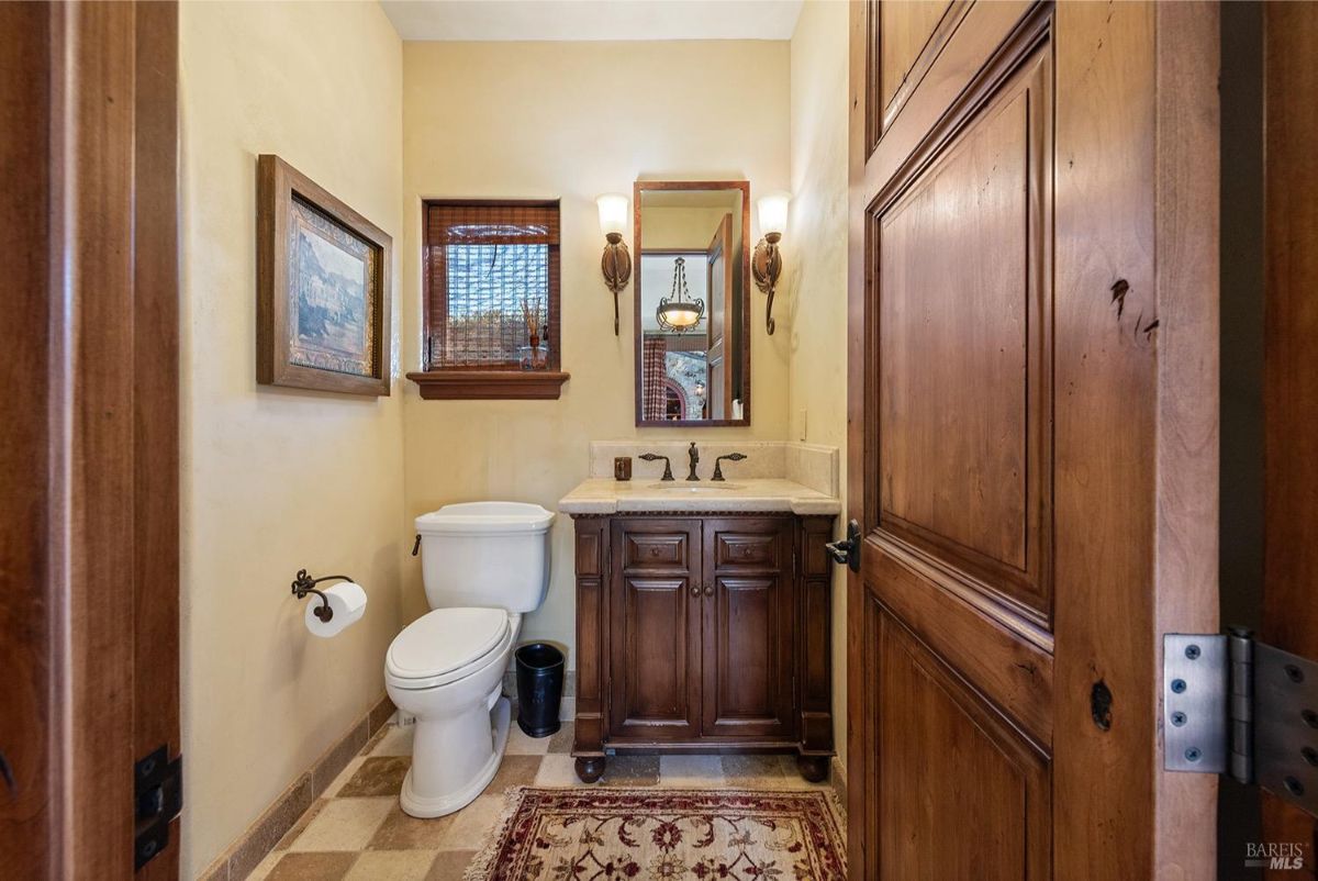 A powder room featuring a single vanity with a wooden cabinet, a rectangular mirror above, a toilet to the side, and two wall sconces on either side of the mirror. The room has a small window with blinds.