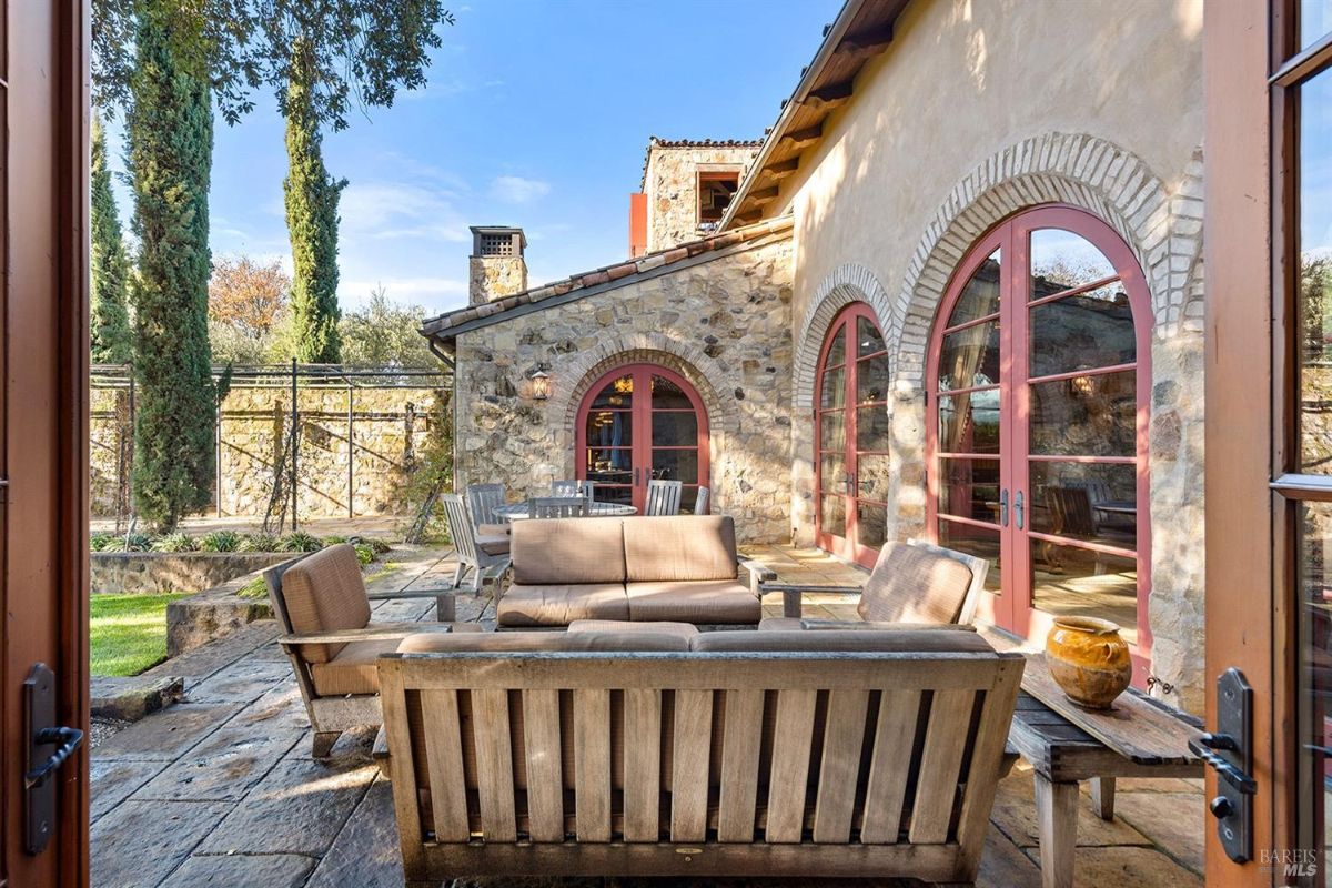 A patio area with wooden furniture, including a sofa and a coffee table, set on a stone surface. The patio leads to arched doors of a stone building.