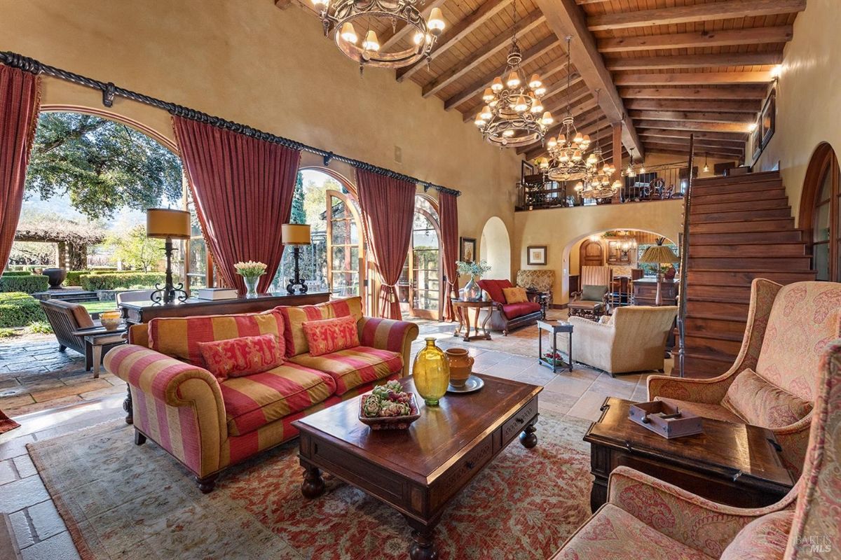 Another perspective of the large room showing seating arrangements including a sofa, chairs, and coffee table. Wooden stairs lead to a mezzanine level, and the arched windows allow natural light to enter the room.