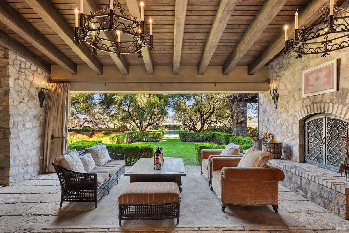 An outdoor lounge area under a wooden ceiling with a stone fireplace, furniture including a coffee table, chairs, and sofas. The view extends to a manicured lawn and trees.