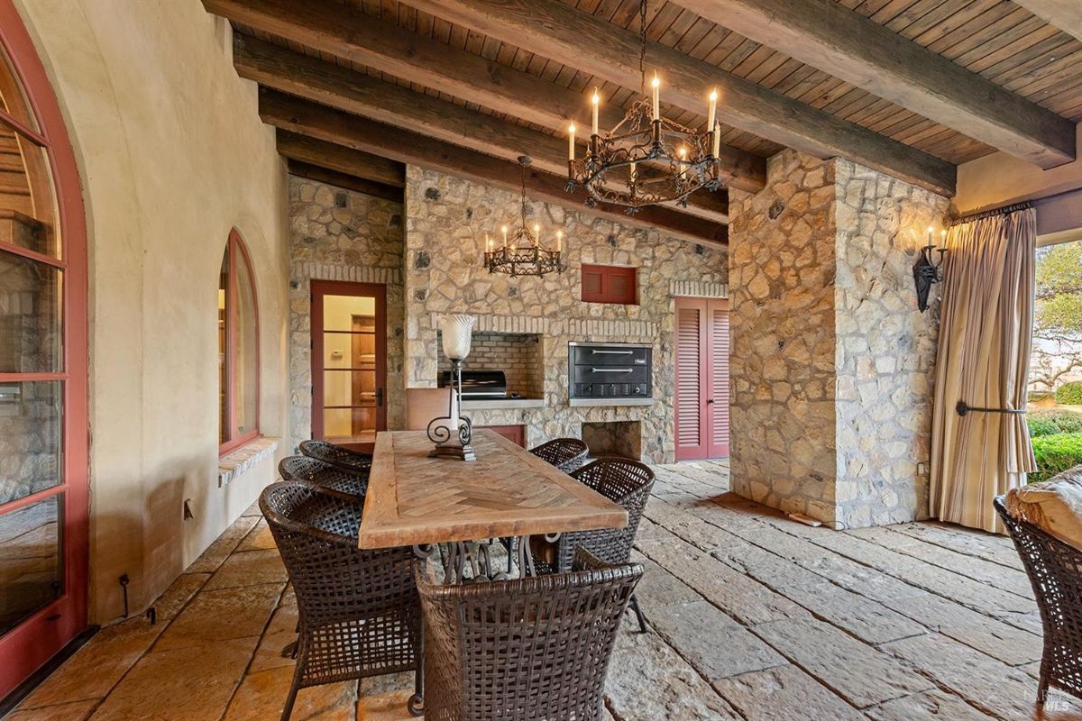 Outdoor covered area with a stone floor, wooden ceiling beams, and chandeliers. Includes a dining table with chairs and built-in cooking appliances.