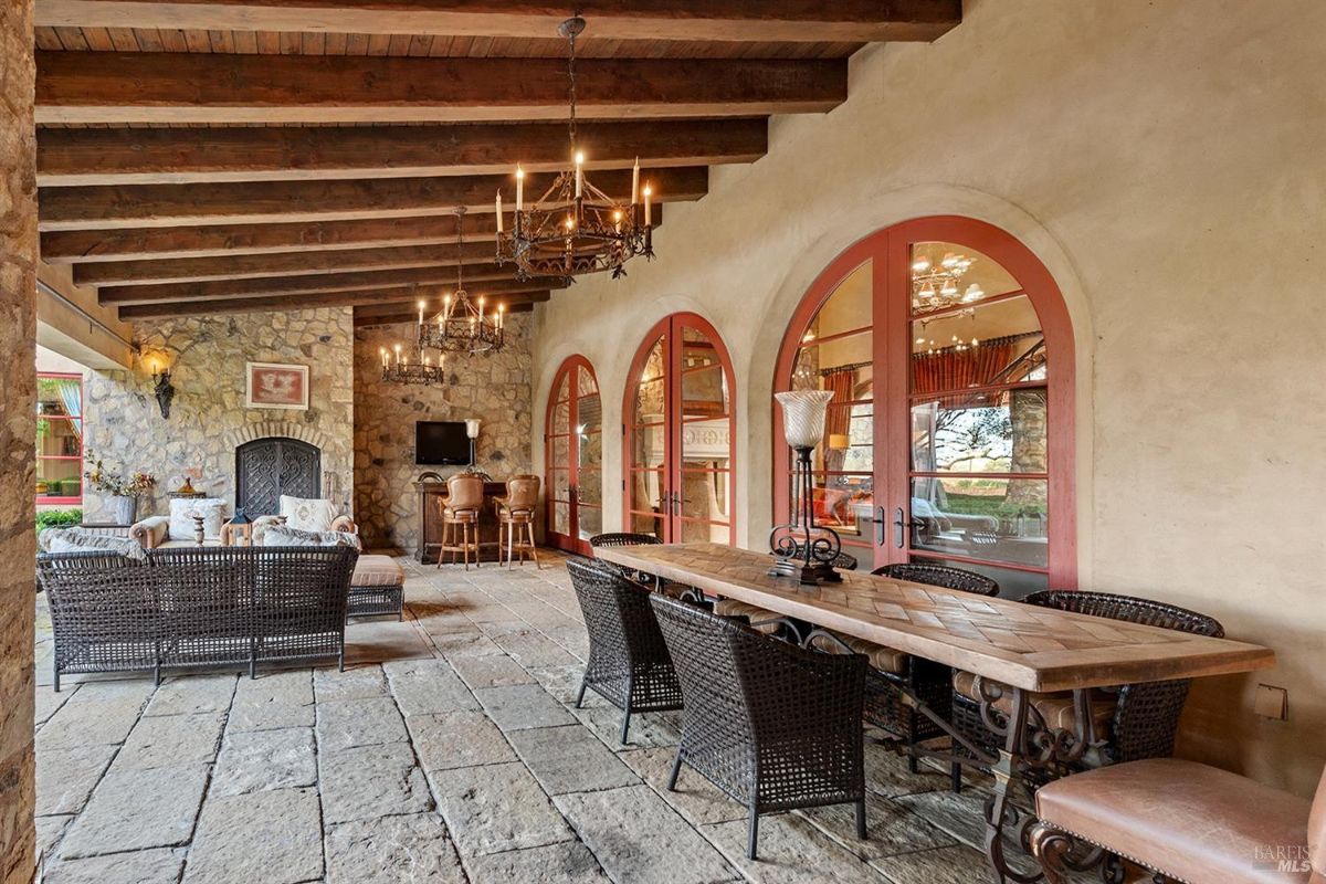 An outdoor covered patio with a stone floor, a long wooden dining table, woven chairs, and arched glass doors leading to the interior.